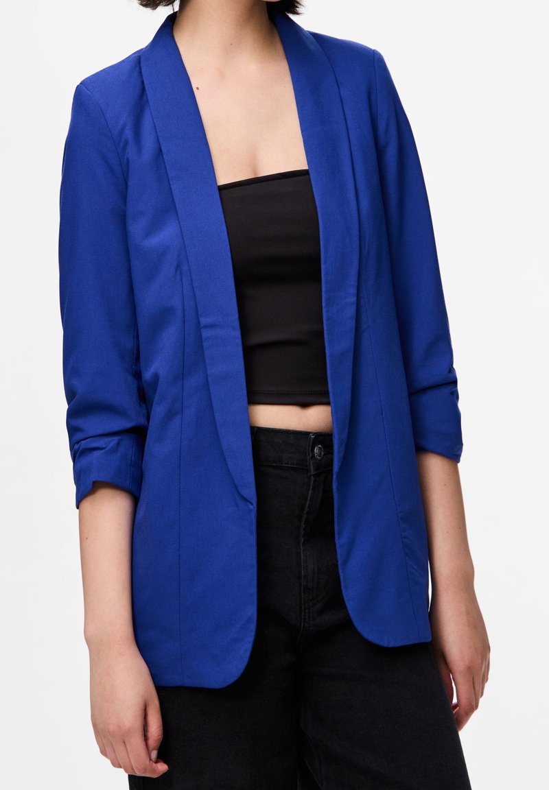 Personne portant un blazer bleu roi à manches retroussées sur un crop top noir sans bretelles et un pantalon noir taille haute sur un fond blanc.