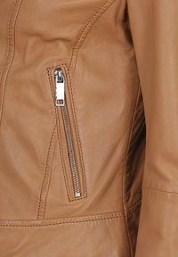 Veste en cuir tan avec une poche zippée diagonale dotée d'une fermeture éclair en métal, texture lisse et couture le long des coutures.