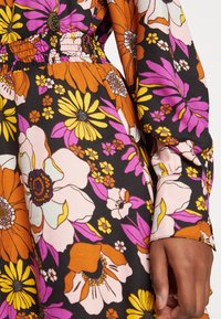Blumenkleid mit schwarzem Hintergrund, großen orangefarbenen, rosa und gelben Blumen. Der Stoff ist glatt; mit gerafftem elastischen Bund. Lange Ärmel.