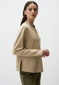 Maglia beige a maniche lunghe con scollo a V e spacchi laterali. Realizzata in tessuto morbido, presenta una tasca sul petto, abbinata a pantaloni verde oliva.