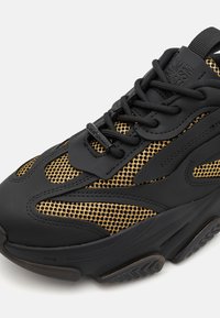 Zapatillas negras y doradas con detalles de malla, suela de goma texturizada y cierre con cordones. Los acentos incluyen paneles de cuero liso y un logotipo.