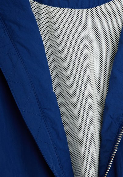 Μπλε μπουφάν με ορατές ραφές, μερικώς ανοιχτό, αποκαλύπτοντας λευκή επένδυση από δίχτυ και μεταλλικό φερμουάρ κατά μήκος της άκρης.