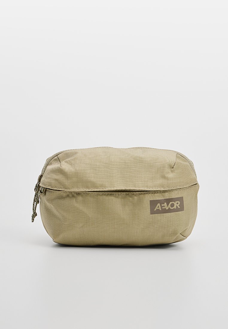 Bolsa de accesorio beige hecha de un tejido duradero. Presenta una forma redondeada con cierre de cremallera y un parche con el logotipo en la parte frontal. Textura suave.