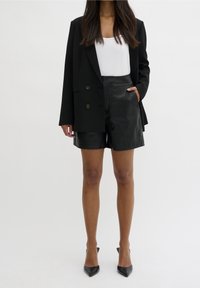 Blazer negro de doble botonadura sobre una camiseta sin mangas blanca, combinado con pantalones cortos de cuero negros de talle alto y tacones negros puntiagudos.