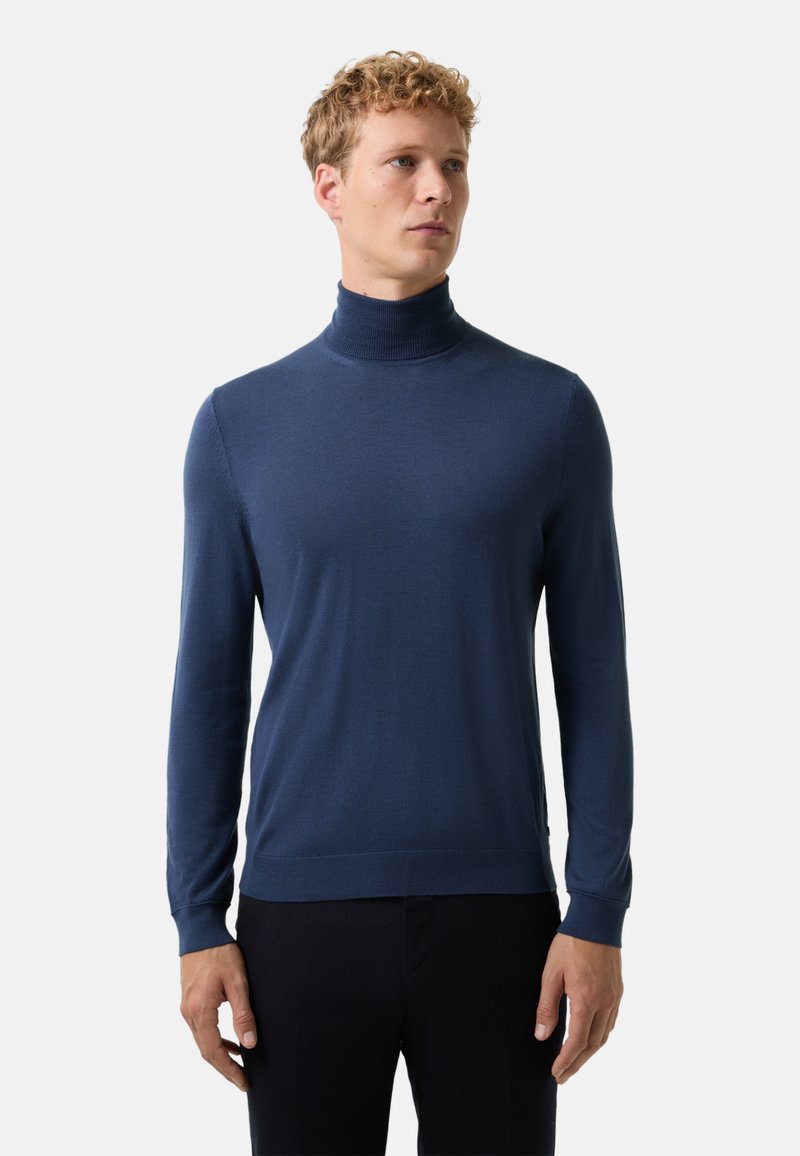 Blonde man in een aansluitende donkerblauwe coltrui en zwarte broek, staand tegen een effen lichte achtergrond.