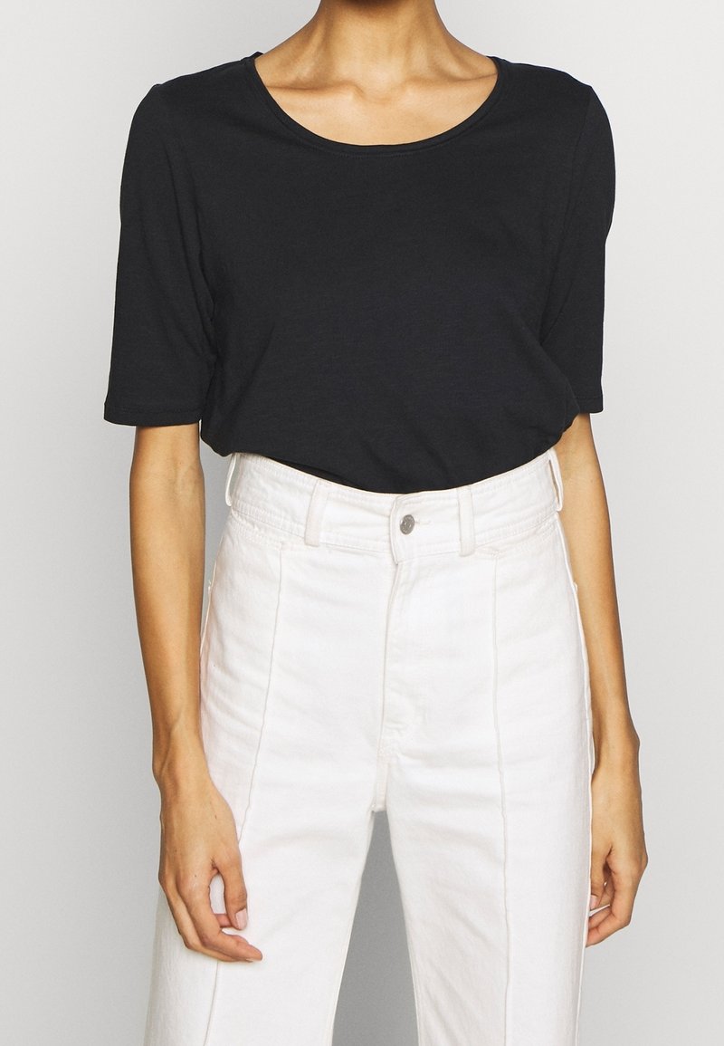 Personne portant un t-shirt noir uni à manches courtes, rentré dans un pantalon blanc taille haute avec un bouton et des passants pour ceinture.