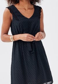 Robe noire sans manches avec un décolleté en V, motif à pois texturé, bordure en dentelle sur les épaules, et une ceinture à nouer à la taille. Tissu léger.
