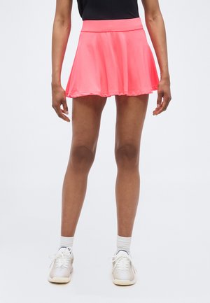 Femeie purtând o fustă evazată roz aprins, top negru, șosete albe și adidași albi, stând într-un decor simplu, cu fundal deschis.