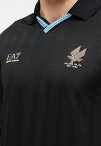 Polo negro con patrón de rayas texturizadas. Detalle en azul claro en el cuello. Logos dorados que muestran la marca y detalles de la fundación.