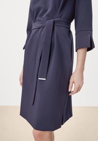 Marineblauwe jurk met een ceintuur in de taille, gemaakt van zachte stof. Kenmerkt zich door driekwart mouwen en een rechte snit met een splitdetail.