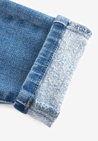 Revers retroussé d'un jean en denim bleu montrant les textures du tissu extérieur et intérieur avec des coutures visibles sur un fond blanc.