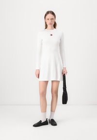 White long-sleeve dress with a slim fit, flared hem, and a small logo detail on the chest. Paired with black loafers and white socks.