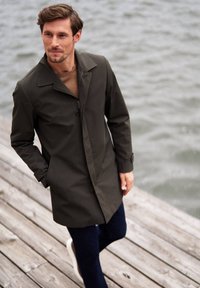 Vihreä vedenpitävä takki, jossa on jämäkkä kaulus, nappisuljenta ja kaksi sivutaskua, yhdistettynä tummiin housuihin ja valkoisiin lenkkikenkään.