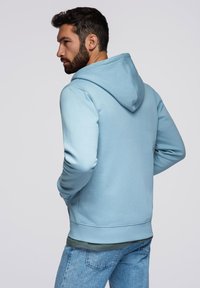 Jasnoniebieska bluza z kapturem i sznurkiem, miękka w dotyku, z prążkowanymi mankietami i pasem. Noszona na ciemnozielonej warstwie, w połączeniu z dżinsami.