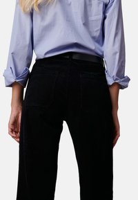 Zwarte corduroy broek met verticale ribbels, voorzien van achterzakken en een riemlus. Gepaard met een lichtblauw shirt met opgerolde mouwen.