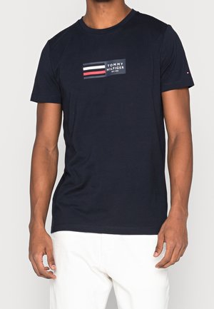 Man draagt een marineblauw Tommy Hilfiger T-shirt met logo en witte, rode en blauwe strepen, gecombineerd met een witte broek tegen een effen achtergrond.