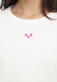 Fehér pamut póló, elöl három apró rózsaszín hímzett szívvel, amelyek háromszög alakban helyezkednek el. Kerek nyakkivágás.