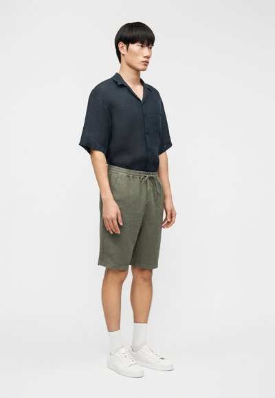 Jeune homme debout portant une chemise sombre à manches courtes avec boutons, un short à cordon de serrage vert olive, des chaussettes blanches et des baskets blanches sur un fond uni.