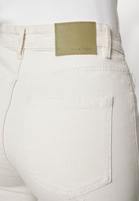 Jeans de ganga brancos com um tecido texturizado, bolsos padrão e um retângulo de pele verde rotulado "DenimProject" na cintura.