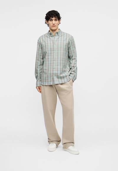 Νέος άνδρας που στέκεται φορώντας πουκάμισο με κουμπιά σε ανοιχτό μπλε και μπεζ καρό σχέδιο, μπεζ παντελόνι με φαρδιά μπατζάκια και λευκά αθλητικά παπούτσια σε απλό φόντο.