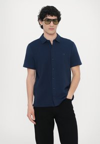 Short-sleeve navy polo shirt with a collar, button front, and a subtle logo patch on the left chest. Worn with black pants and sunglasses.