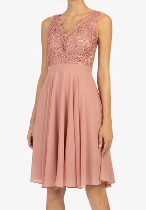 Ärmelloses, staubrosa Kleid mit besticktem Spitzenkorsett und V-Ausschnitt, fließendem, knielangem Chiffonrock an einer stehenden Frau.