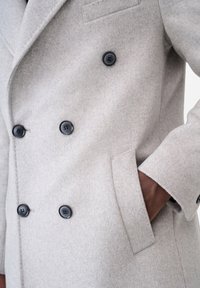 Cappotto doppiopetto in misto lana grigio chiaro, con bottoni in plastica nera, taschino sul petto e tasche laterali, dal taglio sartoriale.