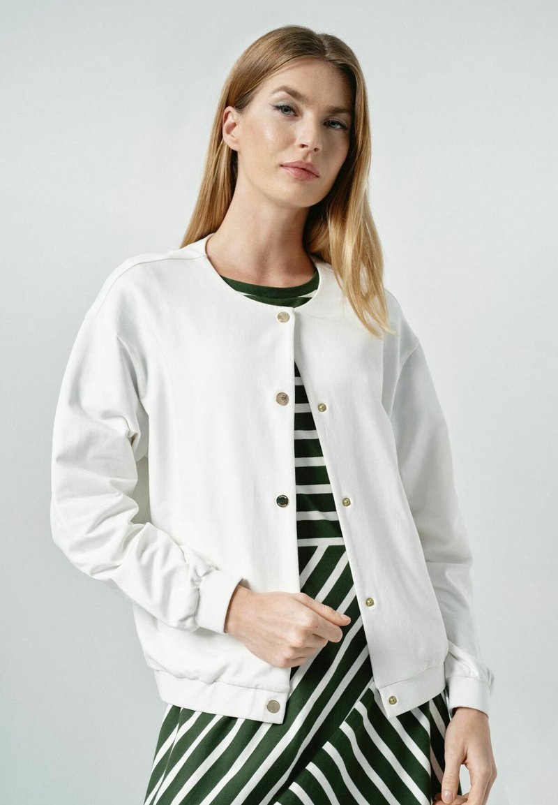 Biała kurtka bomberka z okrągłym dekoltem, złotymi guzikami i elastycznymi mankietami, noszona na zielono-białą sukienkę w paski.