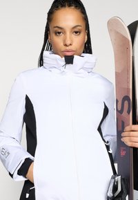 Λευκή και μαύρη σακέτα σκι με ψηλό γιακά, κλείσιμο με φερμουάρ και υφασμάτινες πλευρικές επιφάνειες. Το μοντέλο κρατάει σκι με ροζ λεπτομέρειες.