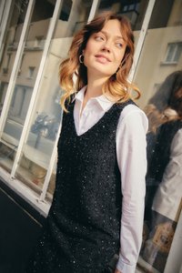 Jeune femme aux cheveux ondulés mi-longs, portant une chemise blanche et une robe noire sans manches à sequins, appuyée contre une fenêtre en verre.