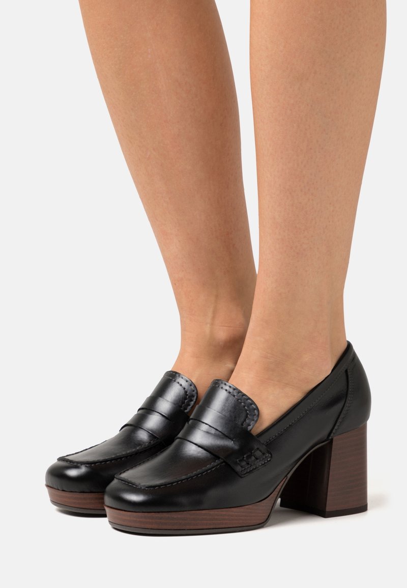Des mocassins à talons blocs en cuir noir portés, avec une finition brillante et une semelle en bois, présentés sur des jambes nues sur fond blanc.