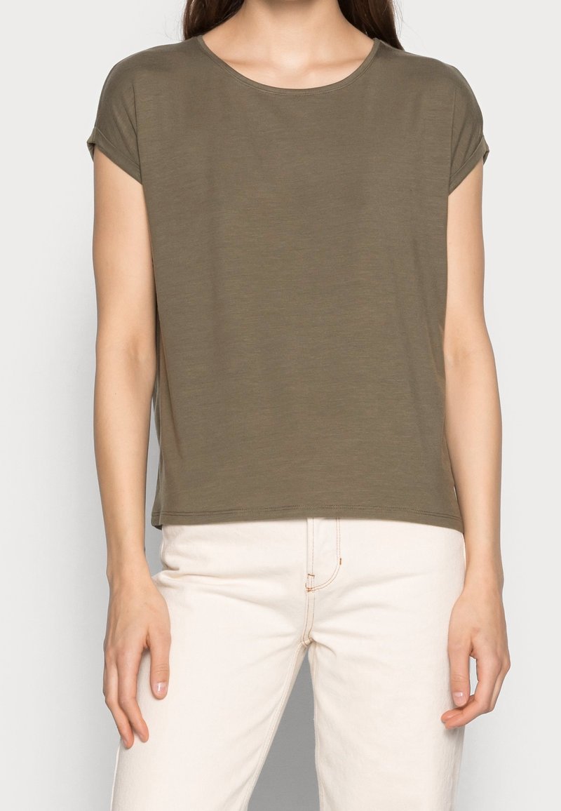 Femme portant un t-shirt à manches courtes vert olive et un pantalon écru, debout devant un fond clair uni.
