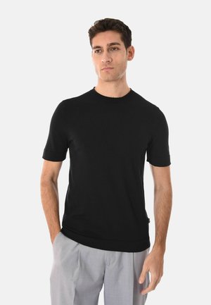 Uomo con capelli corti e scuri che indossa una maglietta nera a maniche corte e pantaloni grigio chiaro, in piedi con una mano in tasca.