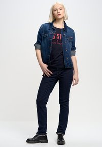 Denim jakna z gumbnim zapiranjem, modre barve, zavihani rokavi. Črna majica s rdečim napisom. Temni kavbojki in črne čevlje, slim fit dizajn.