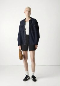 Tamno traper jakna širokog kroja s dva džepa na prsima, bijela majica, crne kratke hlače, bijele čarape i crne tenisice, drži smeđu torbu.