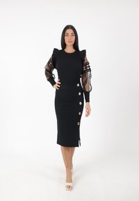 Zwarte jurk met een ribbelige bodice en geraffineerde, doorschijnende kantmouwen. De rok heeft een aansluitend ontwerp en decoratieve gouden knopen aan de zijkant.