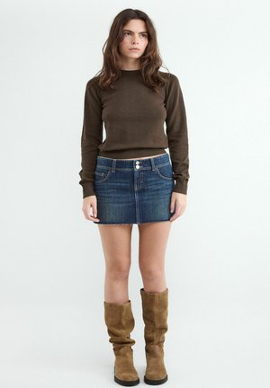 Mladá žena s dlhými hnedými vlasmi, oblečená v hnedý top s dlhým rukávom, modrú rifľovú minisukňu a béžové semišové čižmy po kolená.