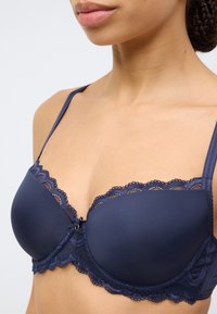 Σουτιέν με μπλε σκούρο μπανέλα και δαντέλα κατά μήκος του πάνω άκρου, με μικρό φιόγκο στο κέντρο και ρυθμιζόμενες τιράντες. Ομαλή υφή υφάσματος.