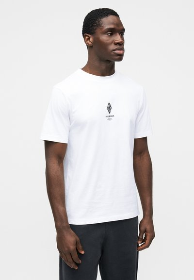 Άσπρο βαμβακερό μπλουζάκι με κοντά μανίκια, με μαύρο λογότυπο στο κέντρο. Το μοντέλο έχει κοντό κούρεμα και ουδέτερη έκφραση.