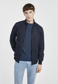 Marynarka bombowa w kolorze granatowym z zapięciem na zamek, dwoma bocznymi kieszeniami, ściągaczami na mankietach i u dołu, noszona na strukturze niebieskiego swetra i jasnoniebieskich dżinsach.