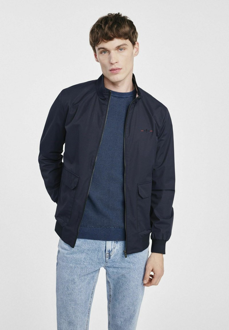 Marynarka bombowa w kolorze granatowym z zapięciem na zamek, dwoma bocznymi kieszeniami, ściągaczami na mankietach i u dołu, noszona na strukturze niebieskiego swetra i jasnoniebieskich dżinsach.