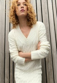 Pulover din tricot cu model căptușit de crem, având un decolteu adânc în V și mâneci lungi cu manșete ușor elasticizate, evidențiind modele texturate pe întreaga suprafață.