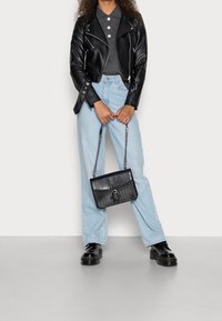 Veste de motard en cuir noir, polo gris, jean large bleu clair et chaussures noires surélevées. Tenant un sac à main noir texturé avec une bandoulière en chaîne.