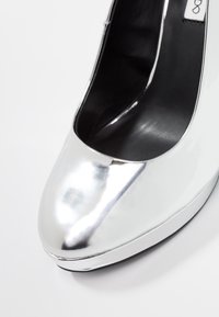 Srebrny but na wysokim obcasie o gładkim, błyszczącym wykończeniu. Okrągły nosek i czarne wnętrze, bez widocznych wzorów lub elementów metalowych.