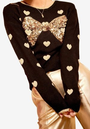 Woman wearing a black sweater with cream hearts and a large gold sequin bow, paired with shiny gold pants and a delicate necklace.