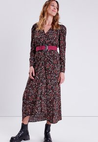 Abito lungo nero con motivo floreale in rosso e crema, caratterizzato da una vita cinchata, maniche a sbuffo e una ampia cintura rosa. Stivaletti neri alla caviglia.