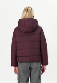 Giacca a vento bordeaux con cappuccio, caratterizzata da un design corto, un motivo a cuciture orizzontali e maniche ampie. Indossata con pantaloni cargo grigi.
