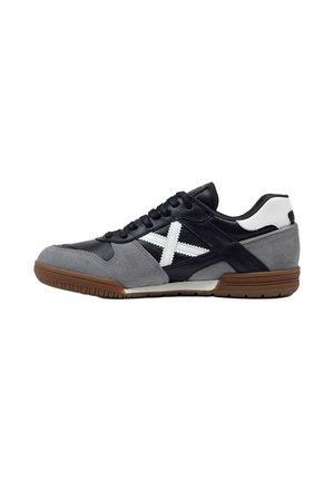 Zapatilla deportiva de perfil bajo con paneles negros, grises y blancos, suela marrón, cordones negros y detalle de cruz cosida en blanco en el lateral.