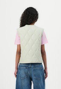Gilet matelassé de couleur vert clair avec un motif en vagues, à manches courtes, porté sur une chemise rose, associé à un jean bleu ample.
