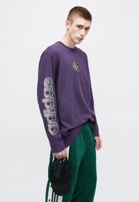 Violeta garām piedurknēm krekls ar apaļu logotipu uz krūtīm un lielu baltu "adidas" uzrakstu uz kreisās piedurknes. Tur zaļas bikses un melna cepure.